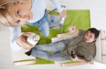 Woman in focus changing light bulb standing on ladder, man in background pointing up from armchair, holding beer, in high angle. Focus placed on light bulb.