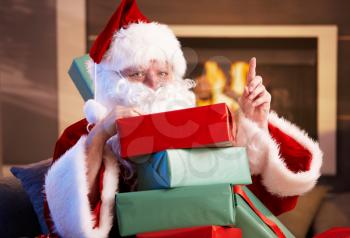 Portrait of Santa Claus sitting by fireplace holding a pile of Christmas presents.