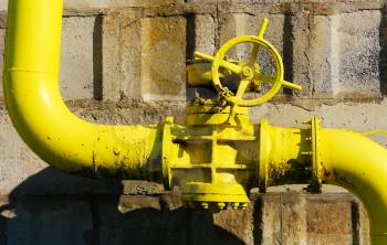 Old gas gate of yellow colour is on a pipe and blocks a pipe. The pipeline with cranes for gas giving on the wall. 