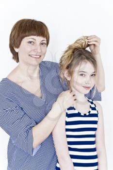 Mother is making hairstyle to her daughter near white wall in striped clothes