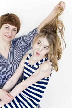 Mother is making hairstyle to her daughter near white wall in striped clothes
