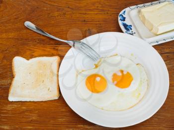 two fried eggs on white plate, fresh toast and dairy butter in butterdish on wooden table