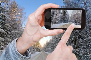 travel concept - tourist taking photo of winter road in snow forest on mobile gadget, Russia