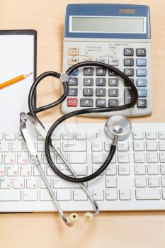 medical still life - phonendoscope on white keyboard, calculator and blank clipboard