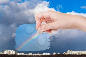 weather concept - hand deletes rainy clouds over city by rubber eraser from image and rainbow and seagull in deep blue sky are appearing