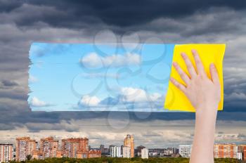 weather concept - hand deletes dark clouds over city by yellow cloth from image and blue sky with white clouds are appearing