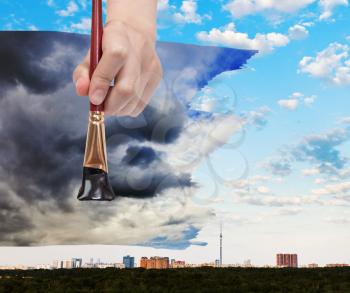 nature concept - seasons and weather changing: hand with paintbrush paints black storm clouds over city