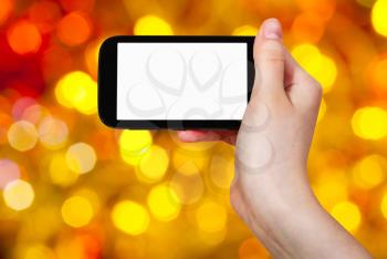 christmas party concept - hand with smartphone with cut out screen on background from brown, yellow and red twinkling Christmas lights bokeh of electric garlands on Xmas tree