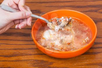 hand holding tablespoon with cabbage soup in meat broth with sour cream over ceramic bowl on wooden table