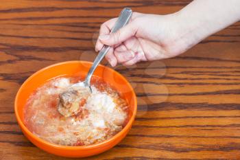 hand holds spoon with cabbage soup in meat broth with sour cream over ceramic bowl on wooden table