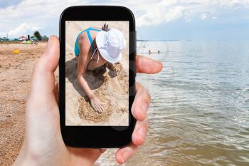 travel concept - tourist photographs girl playing with sand on beach of Azov Sea on smartphone