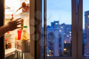 hand reaches for food in refrigerator and view of city through home window in evening