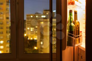 Open door of refrigerator with wine bottles and view of city through home window in night