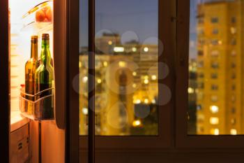 Open door of refrigerator with bottles and view of city through window glass in night