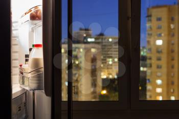 Open door of refrigerator with milk bottles and view of city through window glass in night