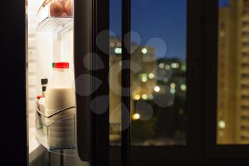 Open door of home fridge with milk bottles and view of city through window in night