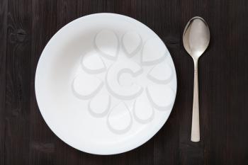 top view of one white deep plate with spoon on dark brown table
