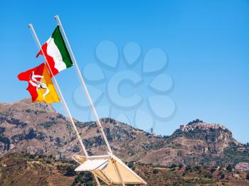 travel to Italy - Italian and Sicilian flags flutter by wind and view of Taormina town on mountain in Sicily