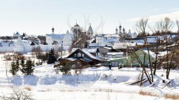 wooden urban houses near walls of Convent of the Intercession (Pokrovsky Monastery) on riverbank of frozen river in Suzdal town in winter in Vladimir oblast of Russia