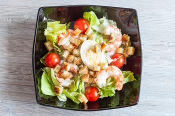 top view of Caesar salad with prawns on black plate on gray wooden table