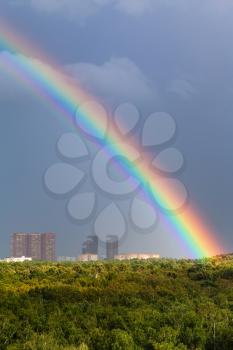 rainbow in gray sky over Moscow city and green trees in Timiryazevskiy urban park in sunny day