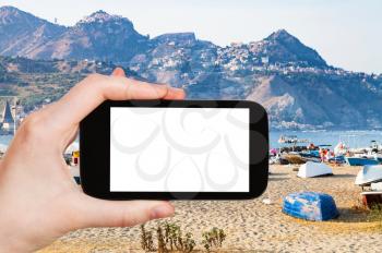 travel concept - tourist photographs urban sand beach and old port in Giardini-Naxos town in Sicily Italy in summer evening on smartphone with cut out screen for advertising logo