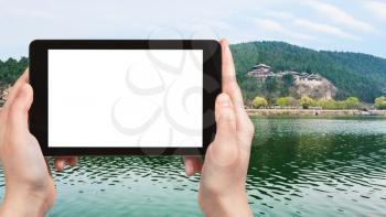 travel concept - tourist photographs East Hill at riverbank of Yi river with Chinese Buddhist monument Longmen Caves (Dragon's Gate Grottoes) on tablet with cut out screen for advertising logo