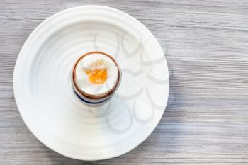 top view of soft-boiled brown egg in cup on white plate on gray wooden board with blank copyspace