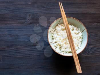 top view of chopsticks above cup with boiled rice on dark brown table with copyspace