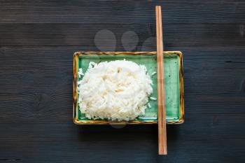 top view of portion of boiled rice and chopsticks on green plate on dark brown table with copyspace