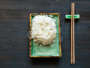 top view of portion of boiled rice on green plate and chopsticks on rest on brown table with copyspace