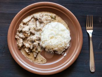 Russian cuisine dish - served portion of Beef Stroganoff (Beef Stroganov, Befstroganov) pieces of stewed meat in sour cream with boiled rice on brown plate on dark wooden board