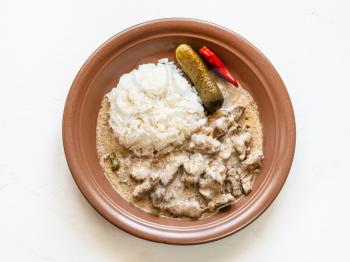 Russian cuisine dish - portion of Beef Stroganoff (Beef Stroganov, Befstroganov) pieces of stewed meat in sour cream with boiled rice on brown plate on white board