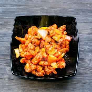 Chinese cuisine dish - top view of black bowl with Fried chicken pieces with pineapple and bell pepper and onion (Sweet and Sour Chicken) on dark wooden board