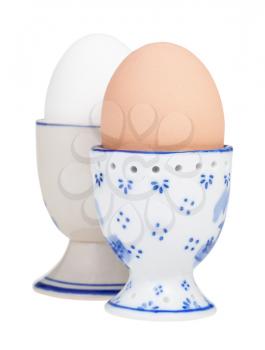 side view of two boiled eggs in ceramic egg cups isolated on white background, the brown egg on foreground