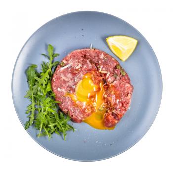 top view of served Steak tartare (raw minced beef meat and chopped onion with raw yolk decorated by fresh arugula salad and lemon) on blue plate isolated on white background