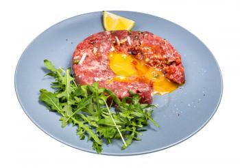 served Steak tartare (raw minced beef meat and chopped onion with raw yolk decorated by fresh arugula salad and lemon) on blue plate isolated on white background