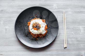 korean cuisine - top view of Kimchi bokkeum bap (fried rice with kimchi, beef and fried egg) on black plate and chopsticks on wooden board