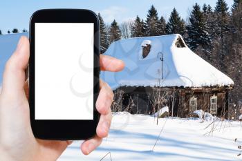 travel concept - tourist photographs of snow-covered old rural houses at the edge of forest in winter in Smolensk region of Russia on smartphone with cut out screen with blank place for advertising