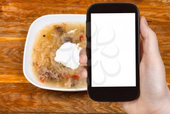travel concept - tourist photographs of russian cabbage soup with stewed sauerkraut and sour cream in ceramic bowl on smartphone with cut out screen with blank place for advertising
