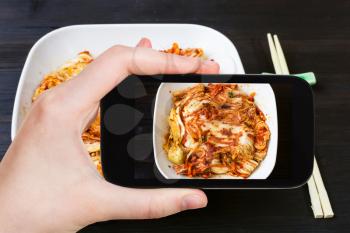 travel concept - visitor photographs of korean cuisine of kimchi appetizer (spicy nappa cabbage) in white bowl on smartphone