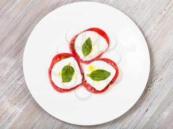 italian cuisine insalata caprese (caprese salad) - top view of stacks from sliced mozzarella cheese, tomato, basil leaf seasoned by olive oil on white plate on wooden table