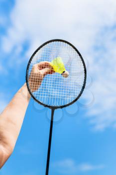 female hand hits yellow shuttlecock by badminton racquet with background from blue sky with white clouds in sunny day (focus on the racquet)