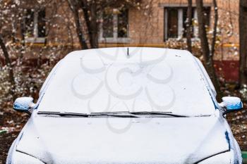 the first snow on car in front of apartment house in snowfall on cold autumn day