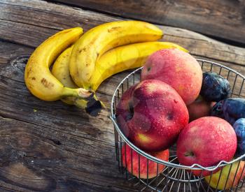 Ripe apples,bananas and plums in metal basket on wooden background