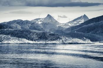 Spring Norwegian landscape with sea coast and mountains with snow. Blue toned monochrome photo, vintage style filter