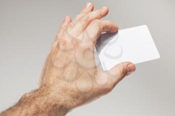 Male hand holds white empty card on gray wall background, close-up photo with selective focus