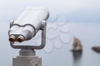 Binocular telescope on a rotating base mounted on an outdoor touristic viewpoint. Close up photo with blurred seascape on a background