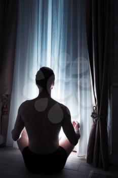 Young adult man is meditating by the window at night