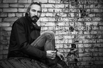 Young bearded man sitting near grungy brick wall, outdoor black and white portrait 
